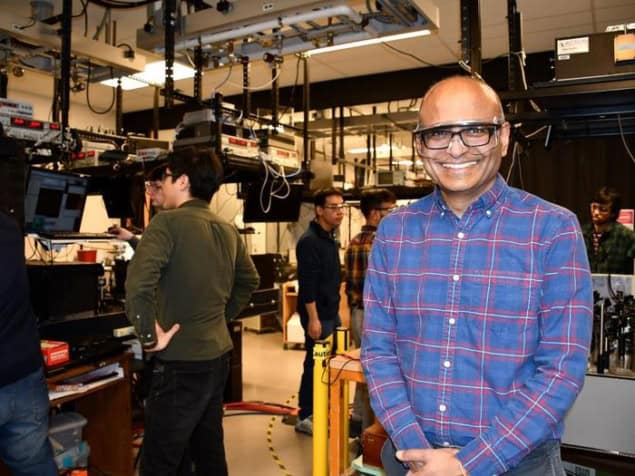 Venkat Gopalan A photo of Venkatraman Gopalan in his optical lab, wearing laser safety goggles and smiling broadly. Other people in the background work at a laboratory bench.